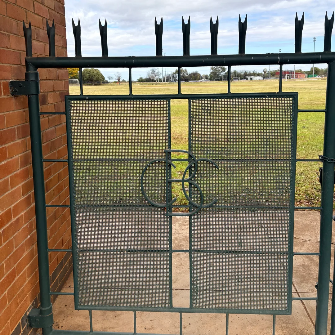 Close-up of a green metal gate at Keeley Memorial Park with the intertwined CBC initials incorporated into the design and the school’s playing fields visible beyond.