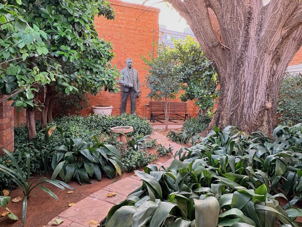 Garden courtyard at the Kimberley Club in Kimberley, with mature trees, planted beds, a bench and a historic statue reportedly relocated from Mahikeng Station.