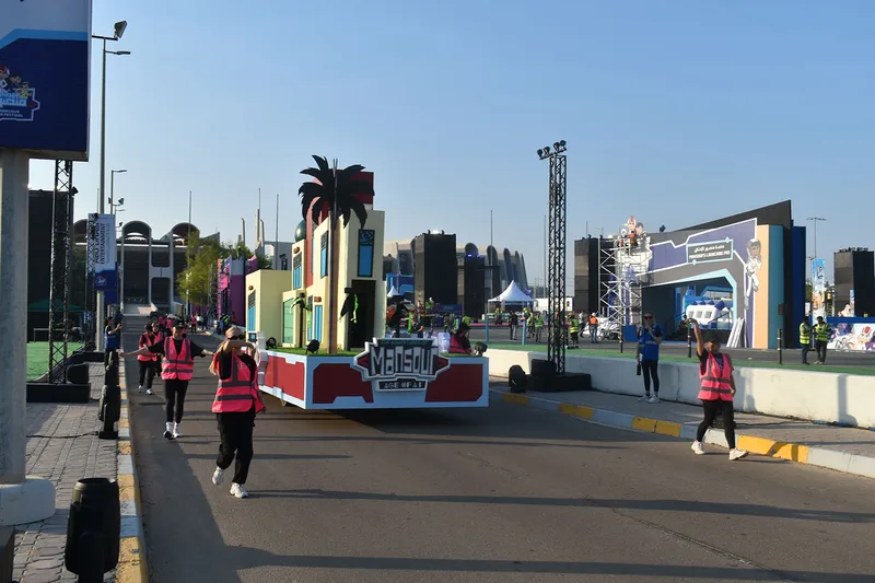 Mansour Festival streetscape float moving through Zayed Sports City with event crew walking alongside.