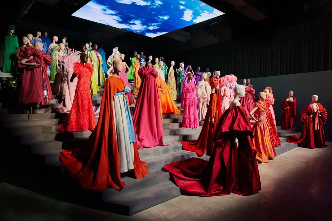 Forever Valentino exhibition in Doha with colourful couture gowns displayed on tiered platforms beneath a large overhead video installation