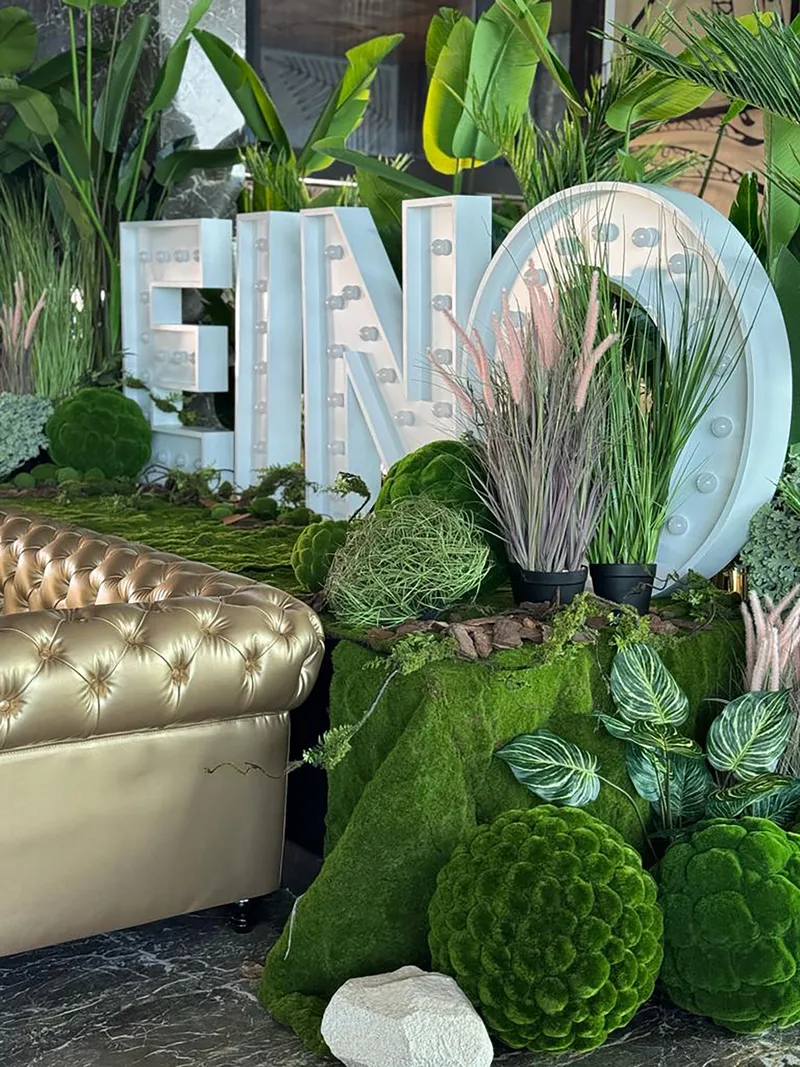 Illuminated EINO marquee letters framed by moss, ornamental grasses and sculptural green foliage.