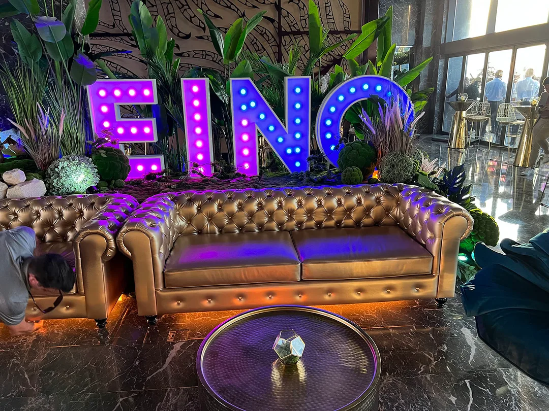 Illuminated EINO marquee letters framed by moss, ornamental grasses and sculptural green foliage.