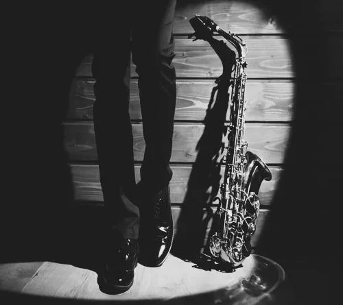 Close up of saxophone in black and white upright against wall next to legs of man. Essex Saxophonist for Hire.