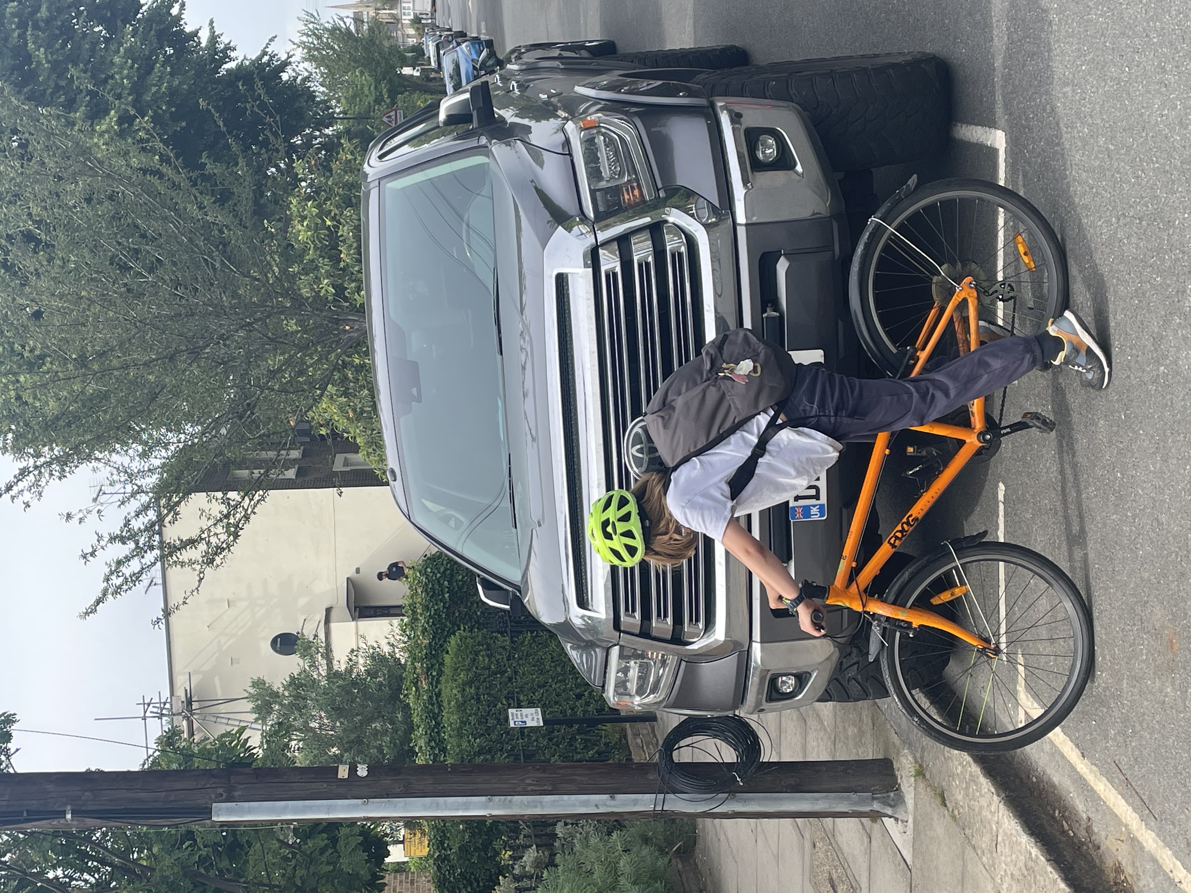 A primary school child on an orange bicycle in front of a huge SUV. The bonnet height is above the child's head and the car width is so big it doesn't fit inside the marked parking bay