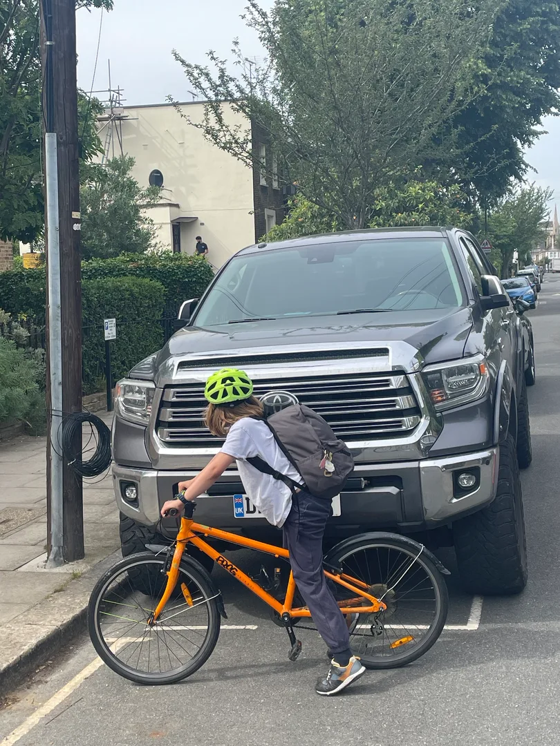 A primary school child on an orange bicycle in front of a huge SUV. The bonnet height is above the child's head and the car width is so big it doesn't fit inside the marked parking bay