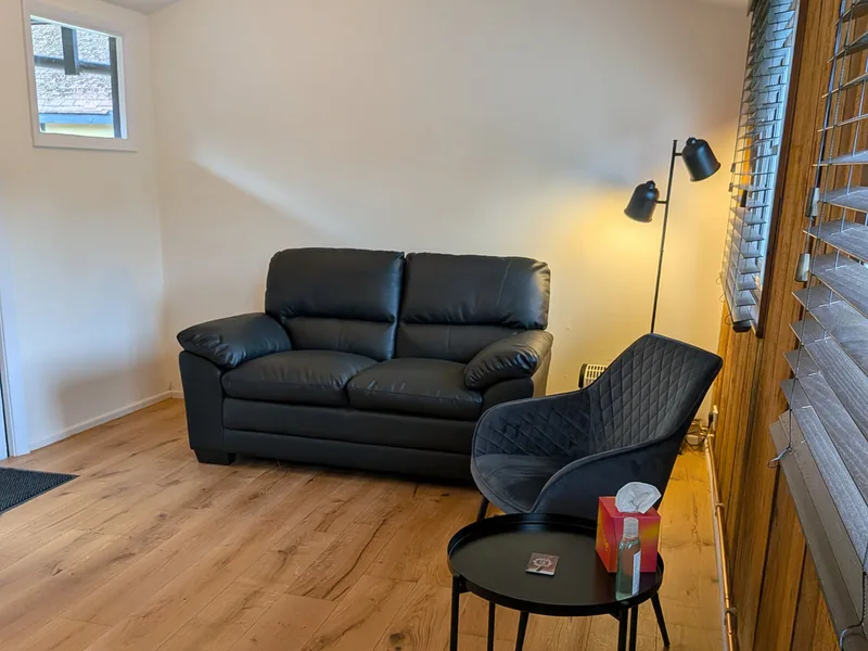 A comfortable, private waiting area at Newhill Osteopathy @ The Stables, Meath Green Lane, Horley with a black leather sofa, soft lighting, and wood-effect flooring for a relaxing atmosphere.
