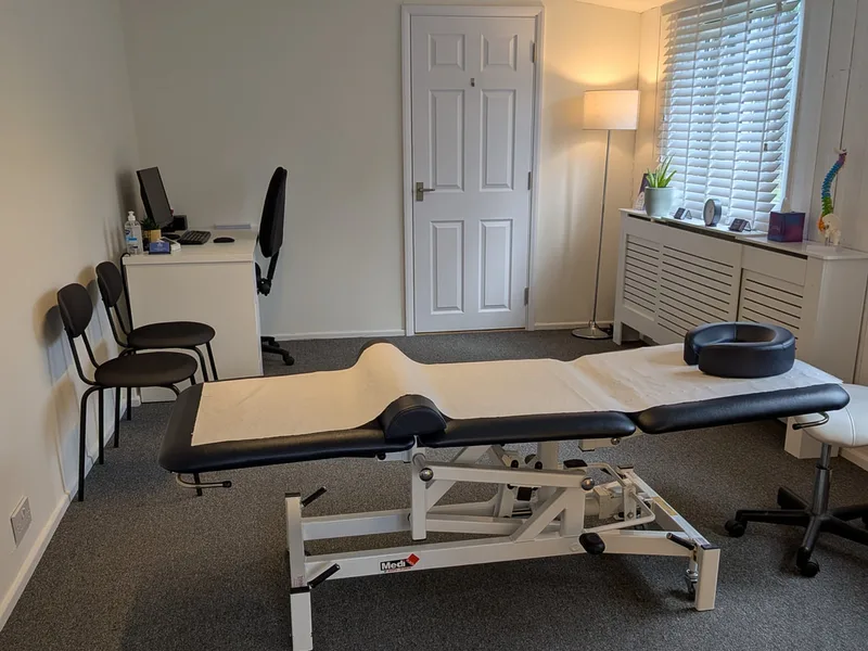 A clean, professional treatment room at Newhill Osteopathy @ The Stables, Meath Green Lane, Horley featuring an adjustable white and black medical couch, desk area, and privacy blinds.