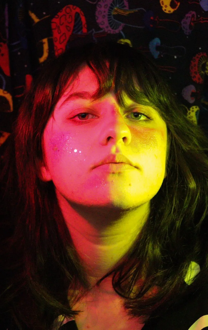 Photo of the artist. She is a woman in her mid to late twenties. The left side of her face has a pink light shining on it, and the right side has a yellow light shining on it.