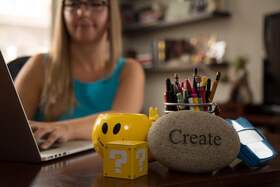 Cassidy sitting at a desk typing on a laptop with 'create' paperweight, a yellow box with question marks, and a yellow smiley face mug in foreground