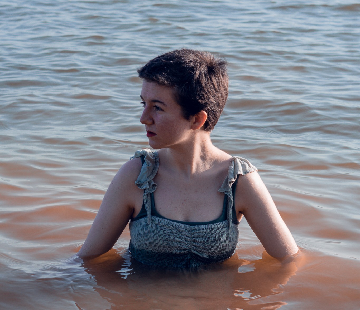 Nadia Radovich in a dress, partially submerged in a lake, looking away from the camera. 