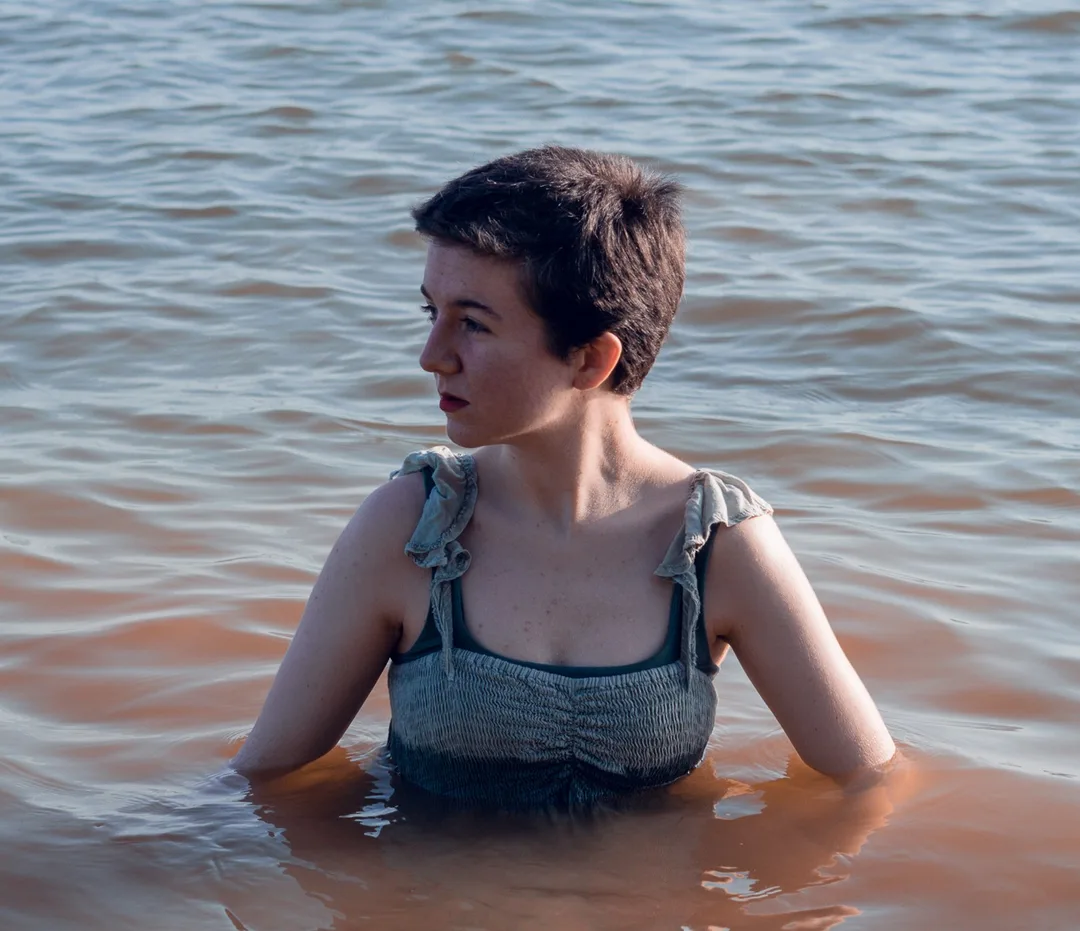 Nadia Radovich in a dress, partially submerged in a lake, looking away from the camera. 
