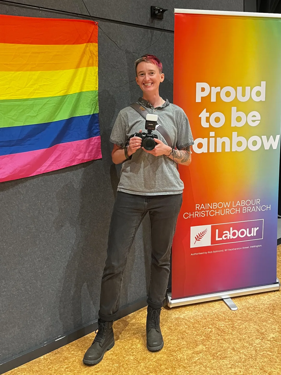 River wears black jeans and a grey shirt, and smiles as they hold up their camera in front of a rainbow pride flag