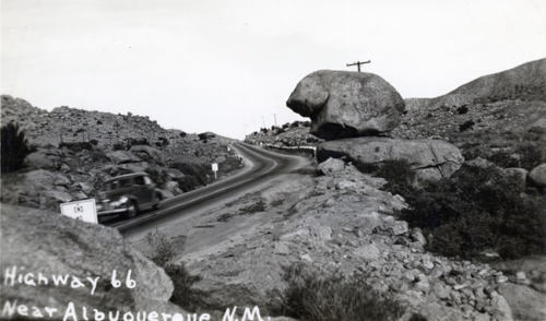 Monochrome photo from behind large rocks next to a two lane road. Written on top: Highway 66 Near Albuquerque N.M.