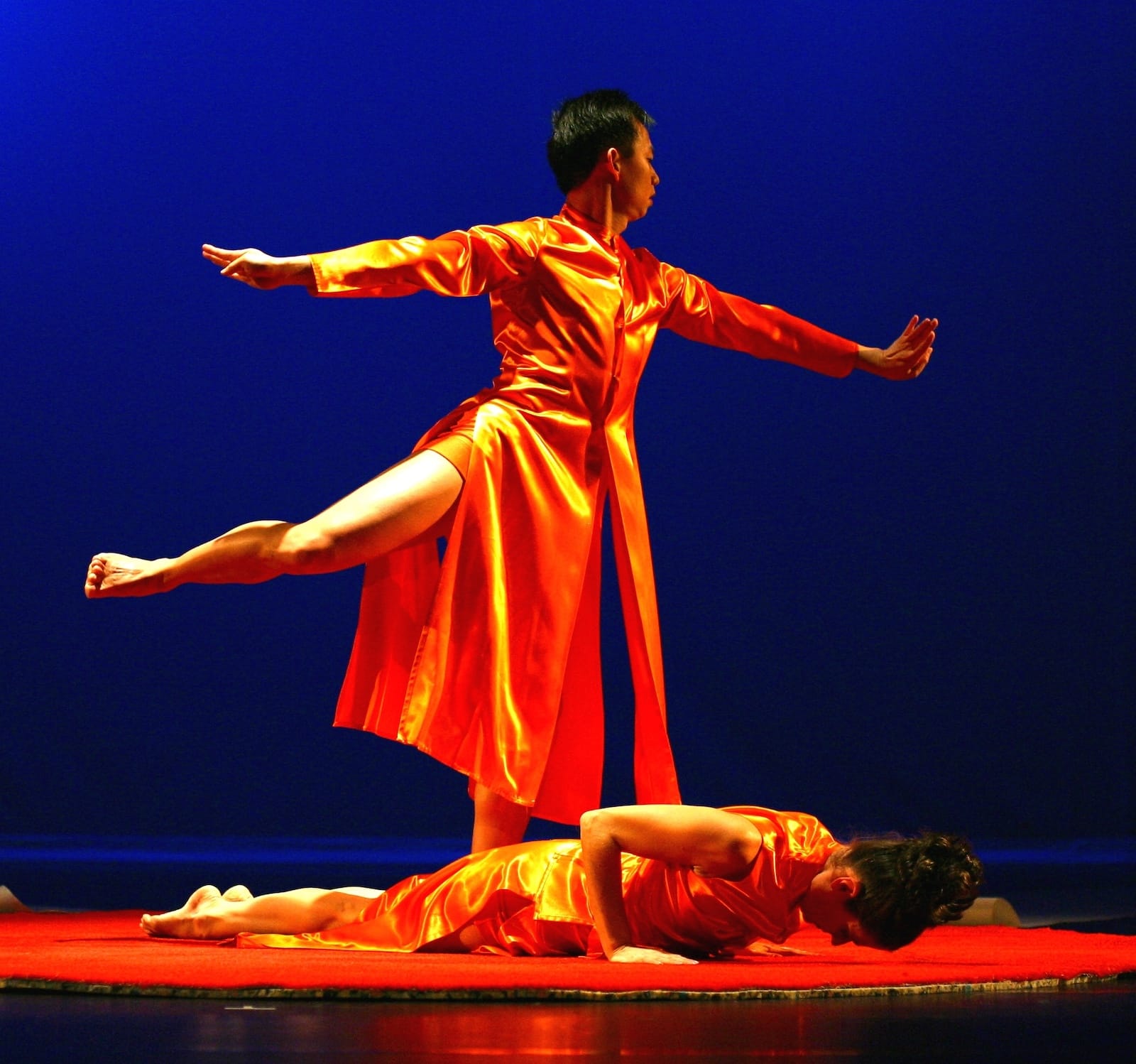 Photo from the show Nocturnal Path. Minh stands on one leg in attitude, wearing a gold robe. Another dancer is below him on the floor.