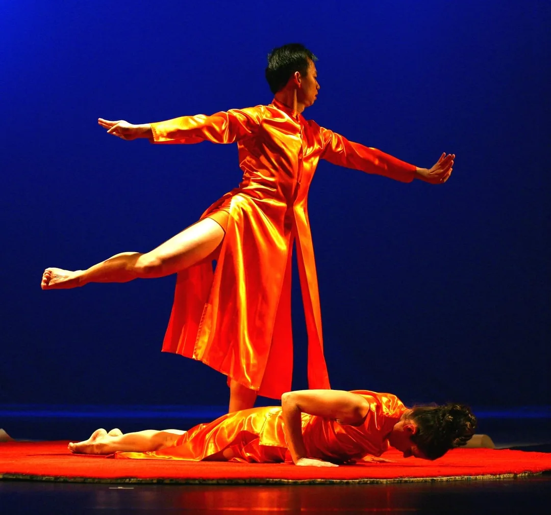 Photo from the show Nocturnal Path. Minh stands on one leg in attitude, wearing a gold robe. Another dancer is below him on the floor.