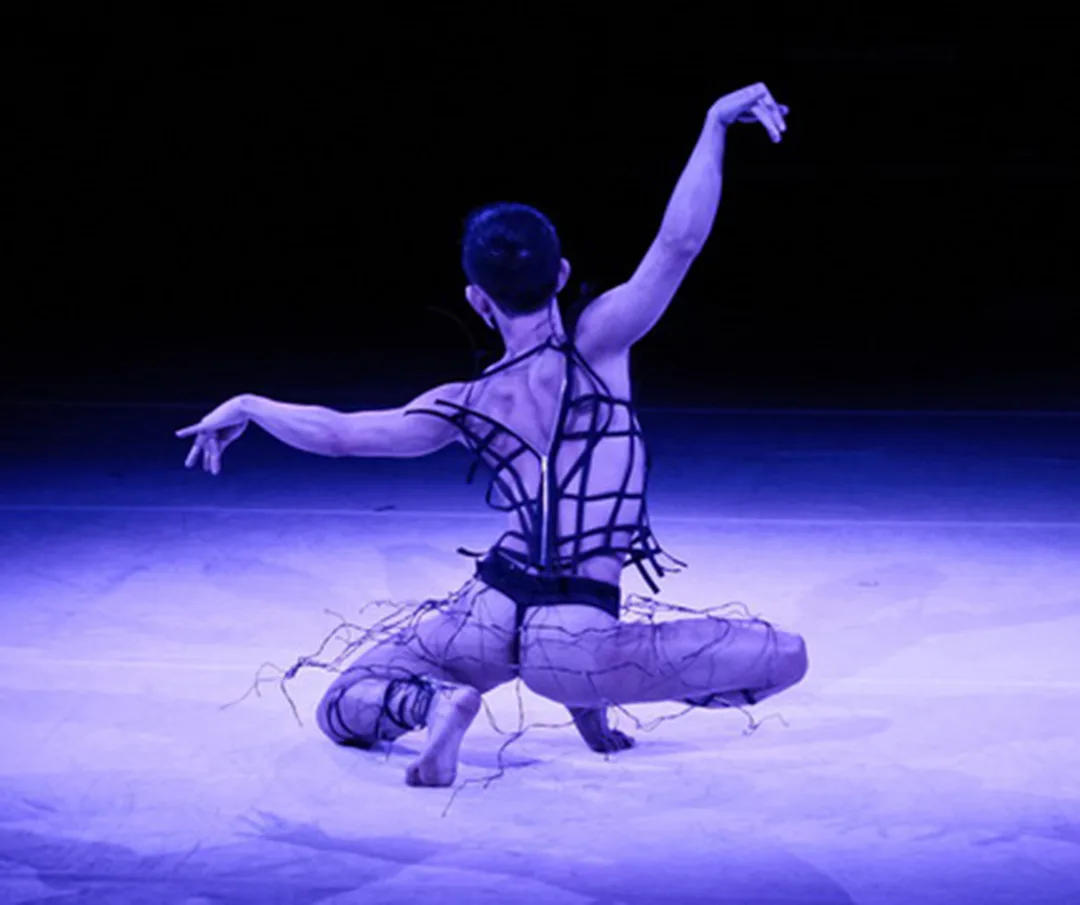 Minh is in a deep toe squat on stage in purple light. His back is to the camera. His outfit shows lots of skin.