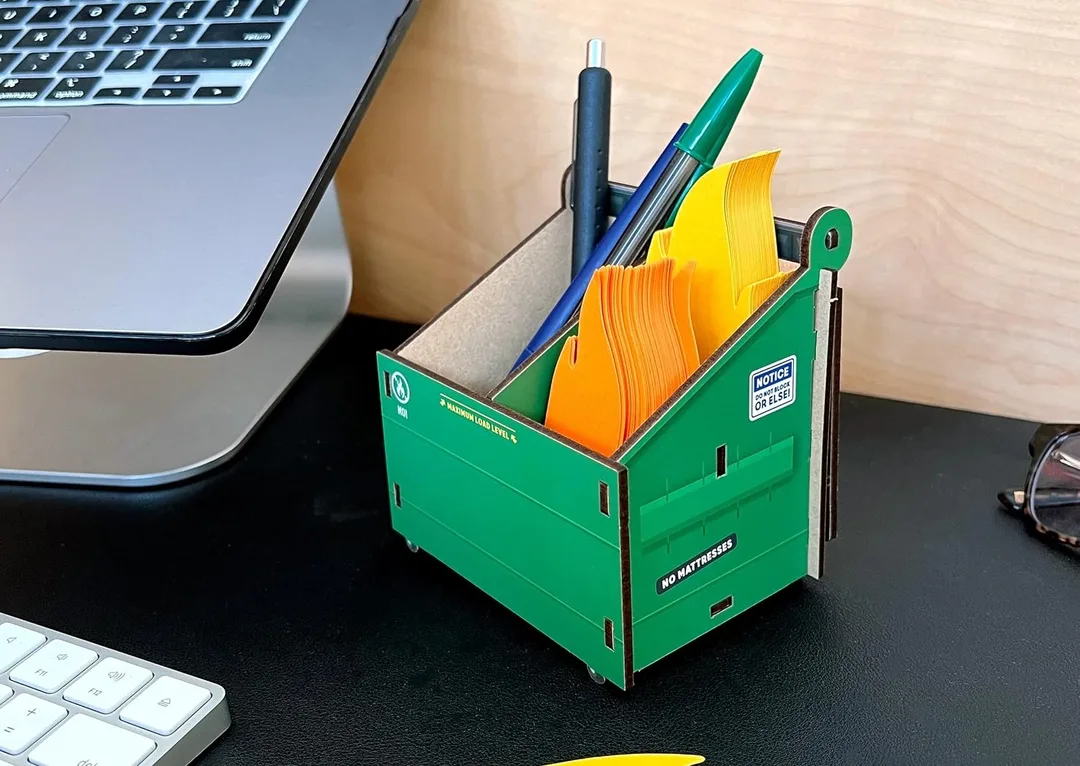 Dumpster shaped pencil holder next to a laptop on a desk.