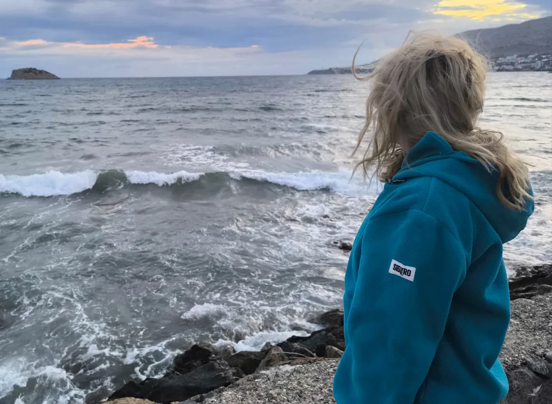 Woman wearing a turquoise SIDURO fleece hoodie beside the sea, with the sleeve label visible.
