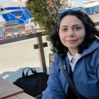 Rose Fazli's headshot. Rose is a woman of South-West Asian descent, sitting at a table in front of a docked ship. She is wearing sunglasses on her head and is smiling at the camera.