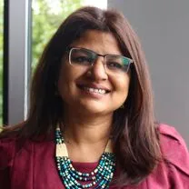 Shazia Shaheen's headshot. Shazia is a woman of South-West Asian descent with long brown hair and glasses. She wears an aquamarine beaded necklace and is smiling at the camera.