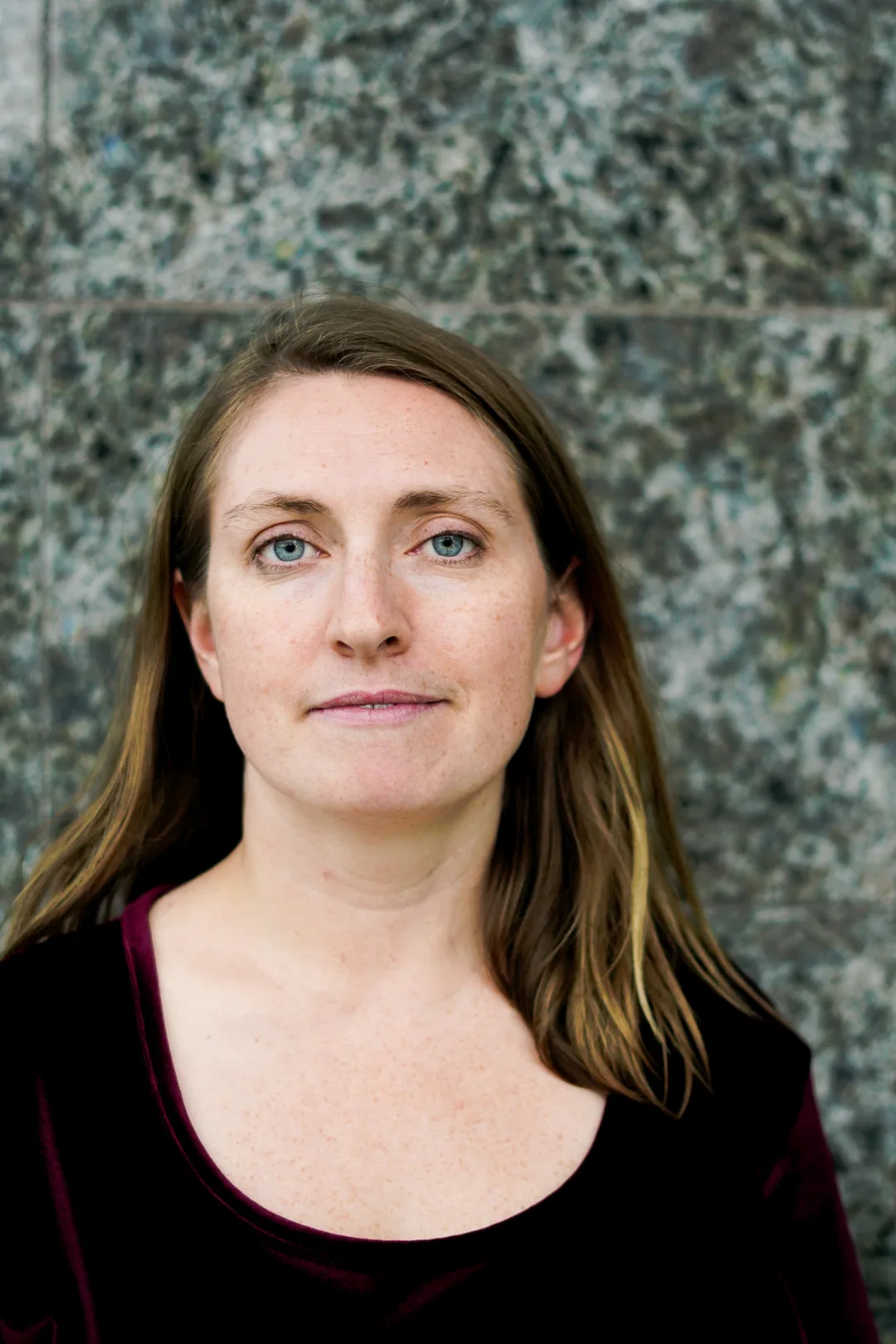 Sharon King-Campbell's is a white woman with long light-brown hair and blue eyes. She is standing against a polished-stone wall and smiling at the camera.