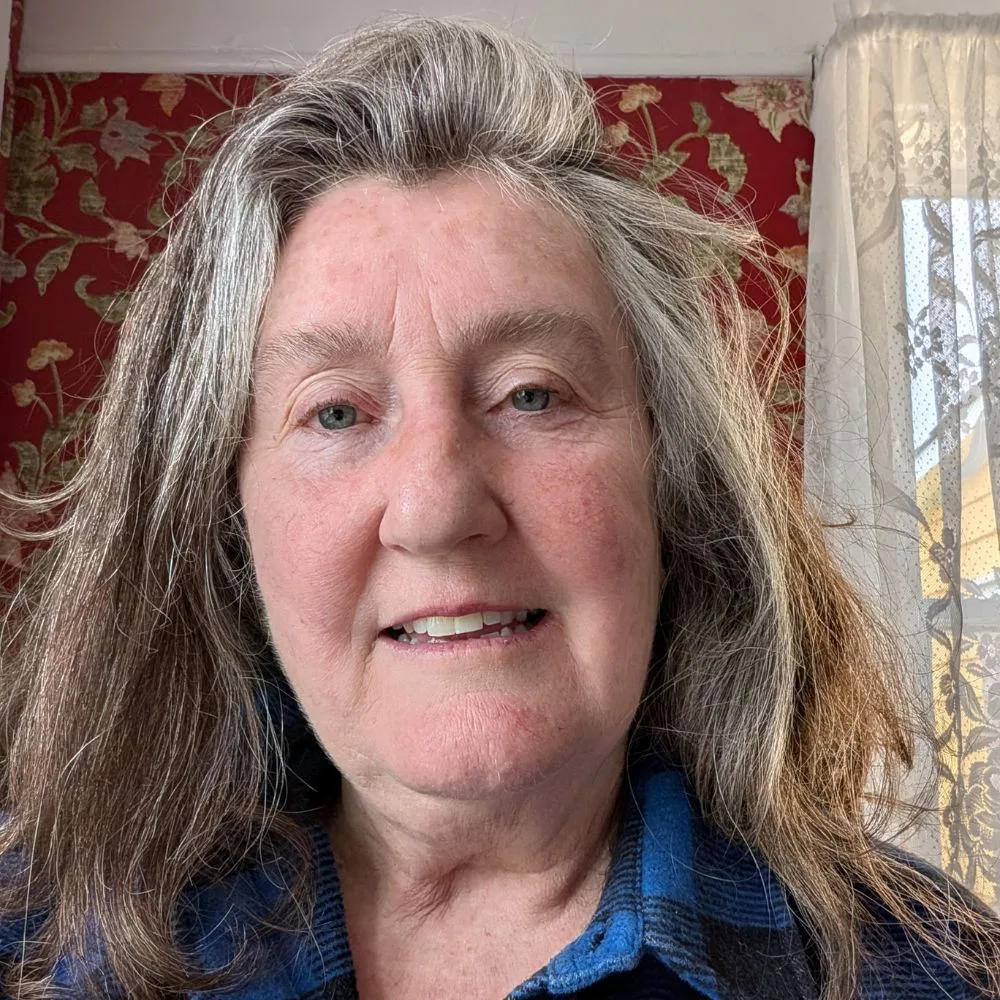 Joan Woodrow's head shot. Joan is a white woman with shoulder-length grey-and-brown hair. She wears a blue plaid shirt and is smiling at the camera.
