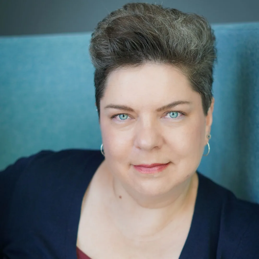 Yuliia Maslak's head shot. She is a white woman with blue eyes and short dark hair, wearing small hoop earrings and smiling at the camera
