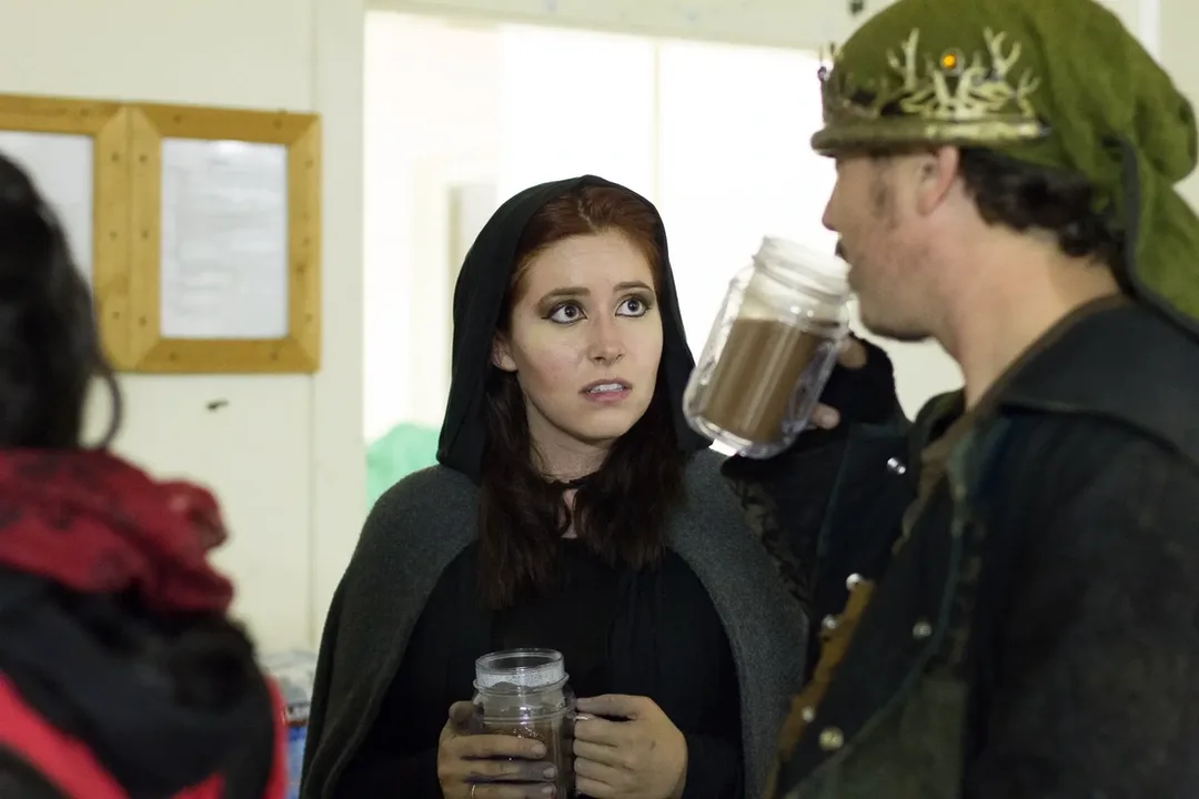 Azrai in a dark hooded cloak stands indoors at a LARP event, holding a large mug and looking attentive and concerned while speaking with another costumed participant wearing a green cap and crown. Both appear in medieval-style clothing, suggesting an in-character conversation.