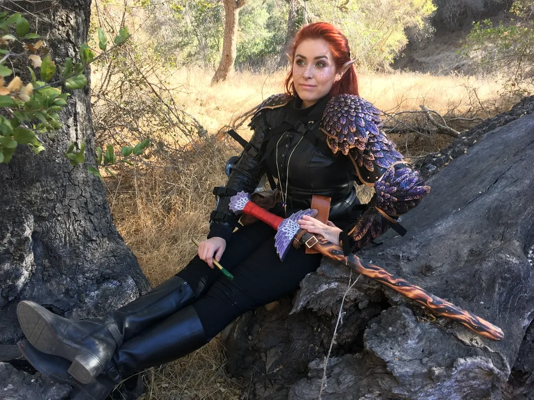 Azrai in detailed fantasy armor with feather sculpted shoulder pieces and elf ears sits on a large tree root outdoors, holding a tin whistle and a flame-decorated sword across their lap. They wear black clothing and tall boots, posing confidently in a wooded landscape.