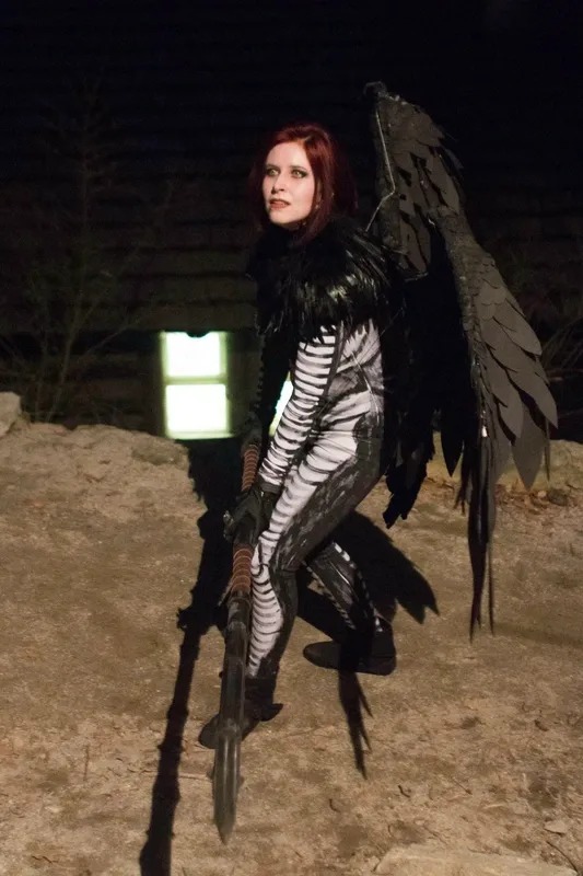 Person in a harpy costume crouches outdoors at night, wearing black feathered wings, a black-and-white patterned bodysuit, gloves, and boots. They hold a long spear and look upward with an alert expression, illuminated by artificial light against a dark background.