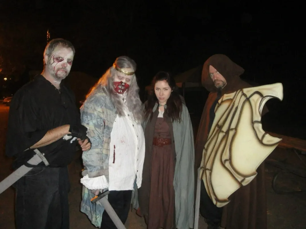 Four people in nighttime fantasy LARP costumes stand together outdoors. Two wear horror-style makeup with pale faces and blood effects, one with long white hair and a gold circlet holding a sword. A hooded figure carries a large bone-like shield, while another person in a cloak and medieval dress stands between them, all facing the camera.