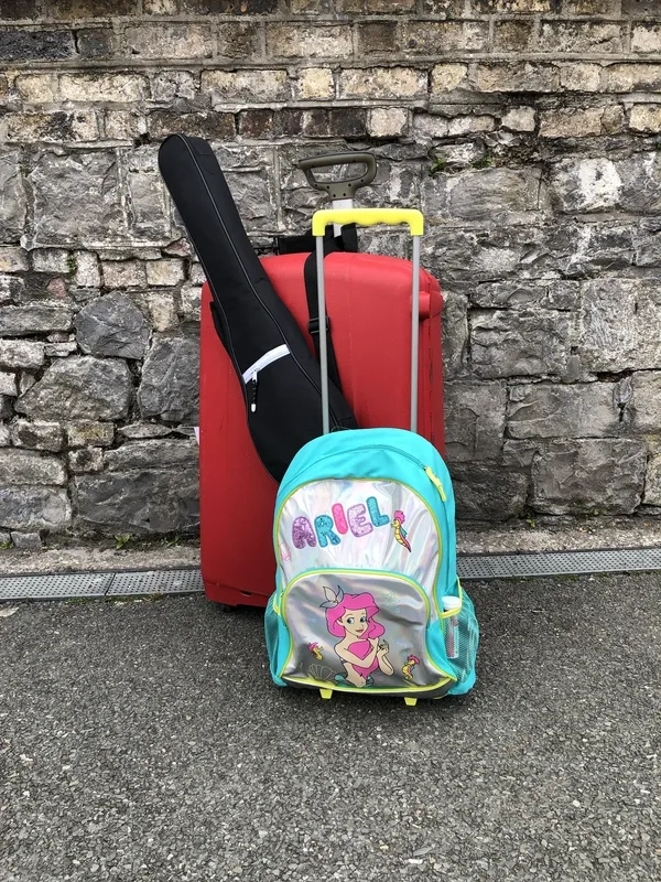 Red hard-shell suitcase, black instrument case, and small rolling backpack with a cartoon mermaid and the name “Ariel” printed on it stand on pavement in front of a rough stone wall, suggesting travel or temporary living out of luggage.