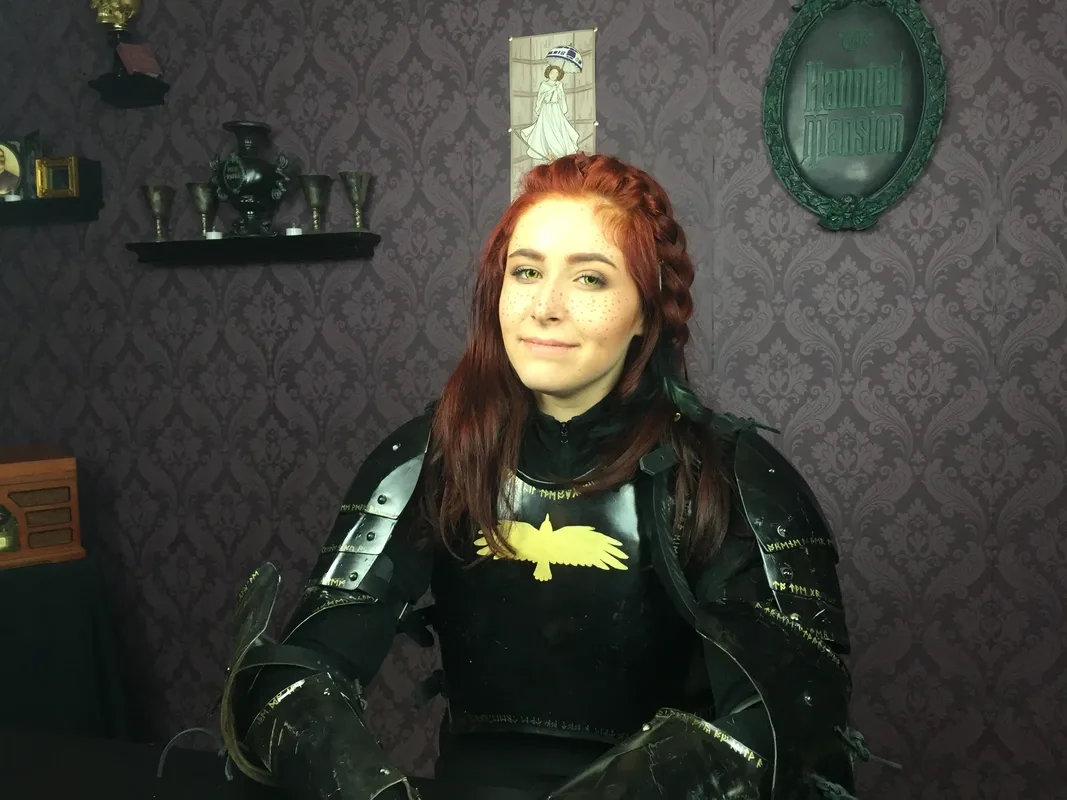 Azrai, with red hair sits facing the camera wearing black fantasy armor with metal shoulder plates and a gold bird emblem on the chest. They are indoors against patterned wallpaper, with decorative shelves and a framed “Haunted Mansion” sign visible behind them.