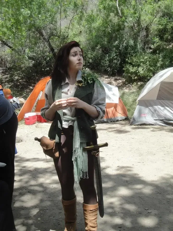 Azrai stands outdoors at a LARP campsite in fantasy medieval-style clothing, including a vest, scarf, boots, and a sword at their belt. Tents and trees are visible behind them as they look off to the side with a thoughtful expression.