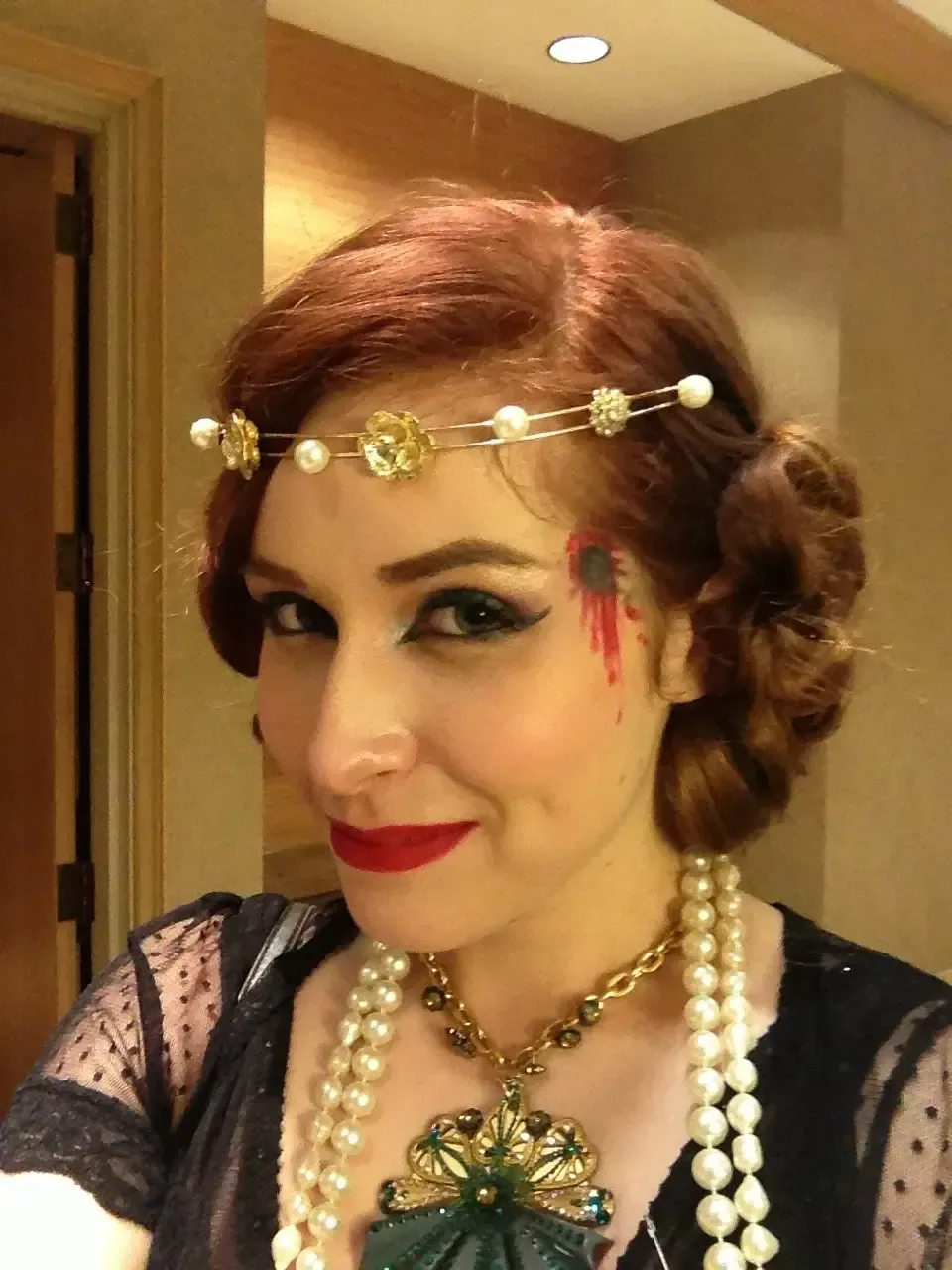Close-up selfie of the same person indoors wearing 1920s-inspired styling: pearl headband, layered pearl necklaces, red lipstick, and finger-wave hairstyle. A theatrical wound-like makeup effect with red and black eyeliner appears at the temple, contrasting with elegant vintage accessories.