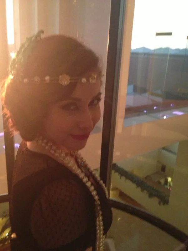 Person in vintage-inspired attire stands beside a large window at sunset, wearing a pearl headband, layered pearl necklaces, and a dark lace dress. Warm golden light silhouettes their face and highlights short, styled hair and classic makeup.