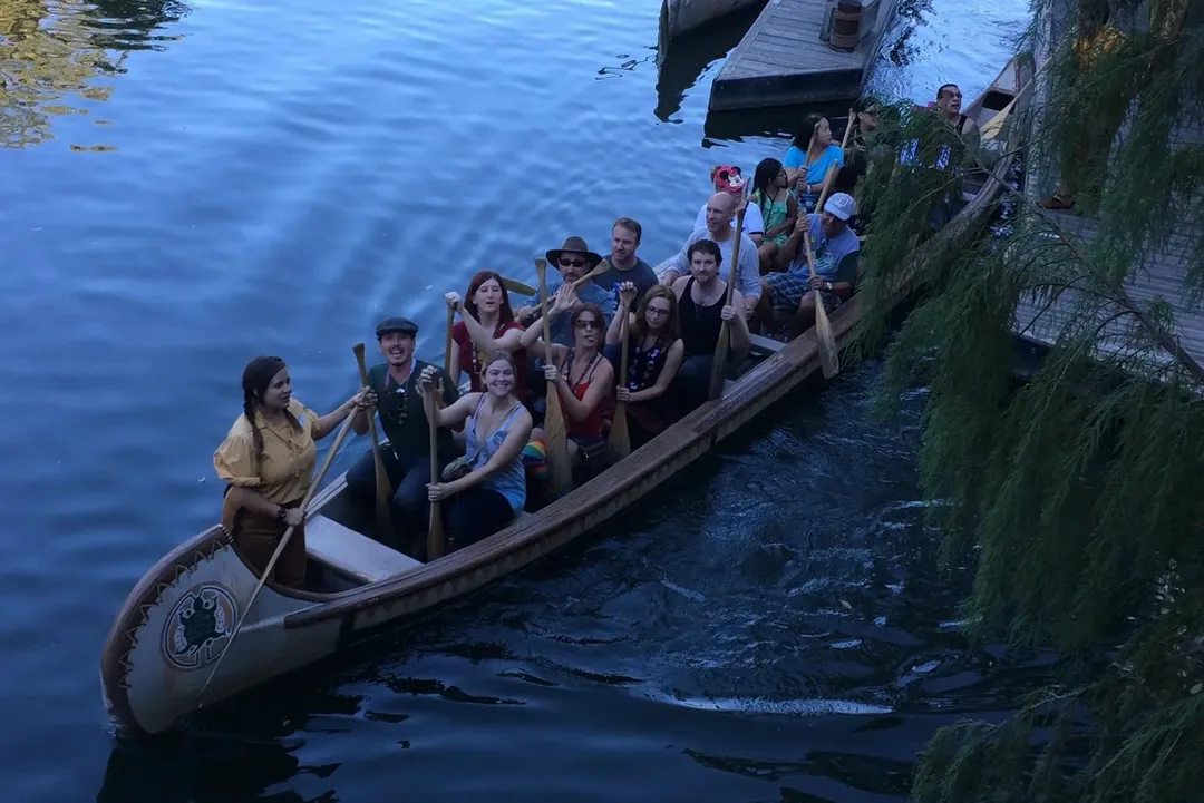 Group of people sit together in a long wooden canoe on calm water near a dock, each holding paddles and looking up toward the camera. Some smile or gesture while the canoe moves forward, surrounded by reflections and overhanging tree branches.