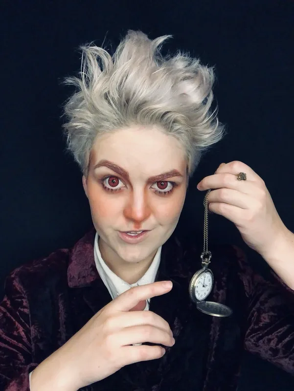 Lou with short, wild platinum-blond hair and theatrical makeup holds up an open pocket watch on a chain, pointing toward it with a knowing expression. They wear a dark velvet jacket against a black background, evoking a mysterious, rabbit-themed character.