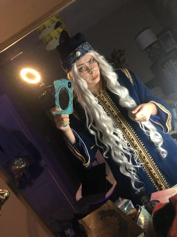 Azrai taking a mirror selfie while wearing a long white wig, round spectacles, and a dark blue robe with gold trim and a tall black hat, resembling a wizard costume. A ring light glows behind them, and books and costume accessories are visible on the table in the foreground.