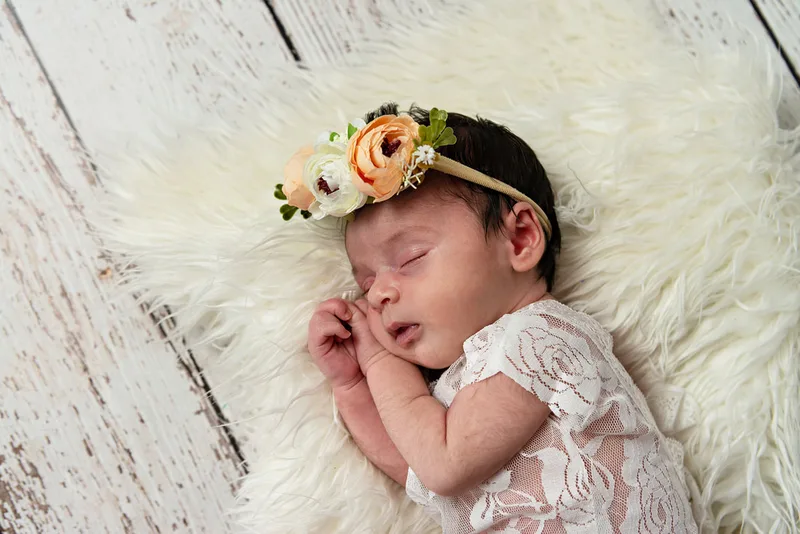 color photo of a baby newborn girl in a white lace outfit with a flower headband laying on a white fur rug
