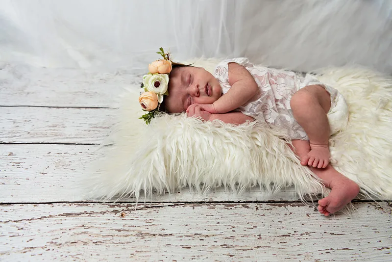 color photo of a baby newborn girl in a white lace outfit laying on a white fur rug with a flower headband