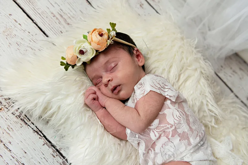 color photo of a baby newborn girl in a white lace outfit with a flower headband laying on a white fur rug