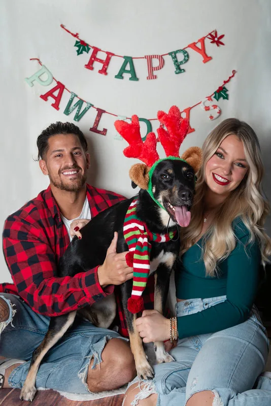 photo of a man and woman and their hound mix dog in front of a Happy Pawlidays banner