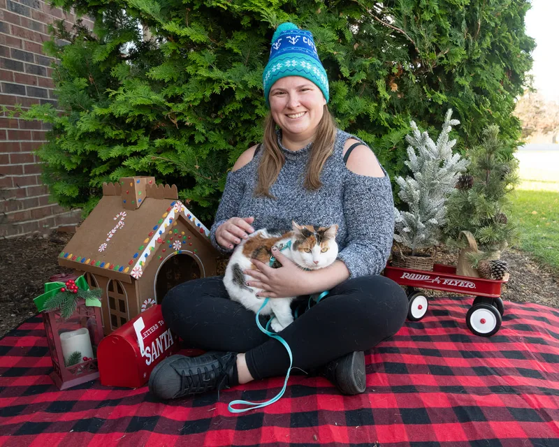 Color photo of a cat and its owner in front of Christmas props on a buffalo plaid blanket at the Bayport-Blue Point Library. The photo was taken by Laura of Laura Pea Photography, a Long Island based pet photographer. 