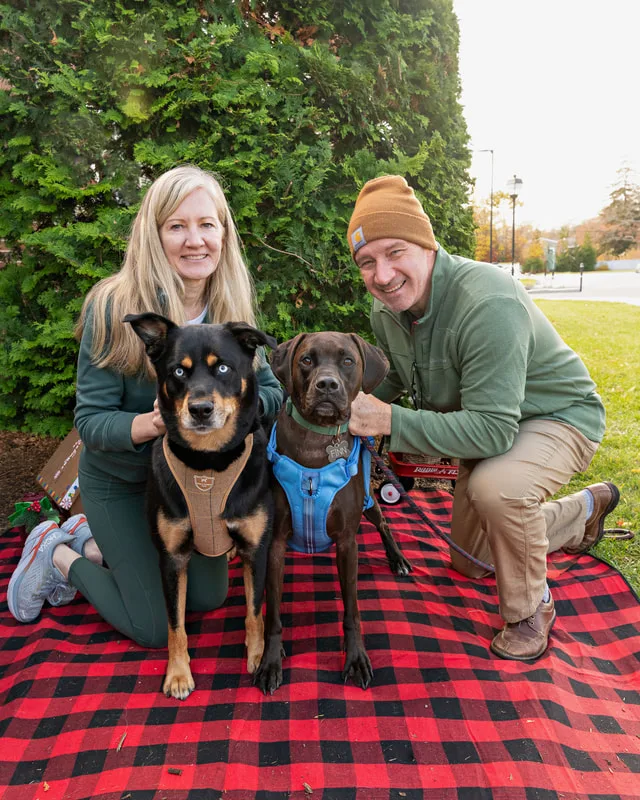 Color photo of two dogs and their owners in front of Christmas props on a buffalo plaid blanket at the Bayport-Blue Point Library. The photo was taken by Laura of Laura Pea Photography, a Long Island based pet photographer. 