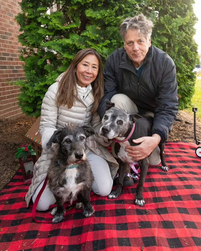 Color photo of two dogs and their owners in front of Christmas props on a buffalo plaid blanket at the Bayport-Blue Point Library. The photo was taken by Laura of Laura Pea Photography, a Long Island based pet photographer. 