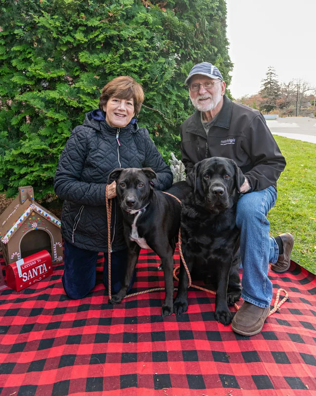 Color photo of two dogs and their owners in front of Christmas props on a buffalo plaid blanket at the Bayport-Blue Point Library. The photo was taken by Laura of Laura Pea Photography, a Long Island based pet photographer. 