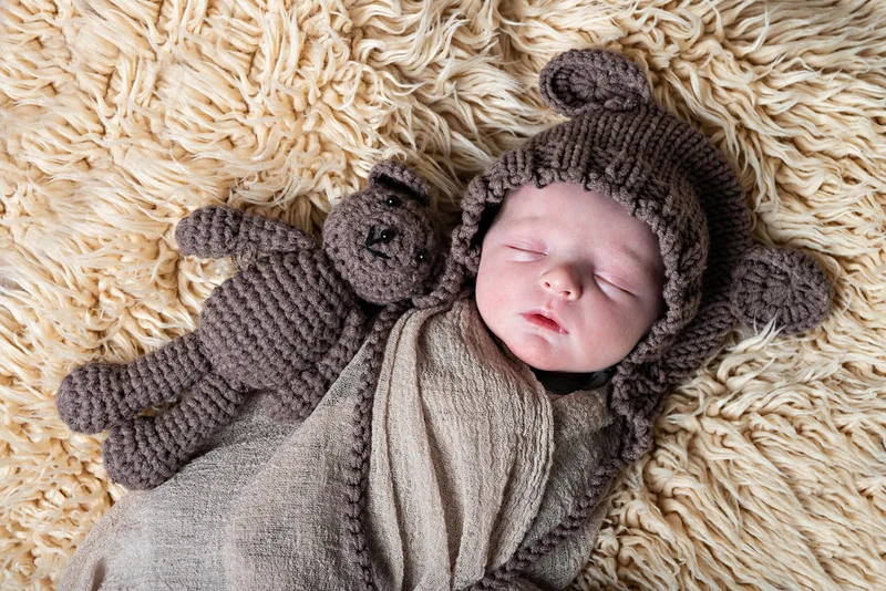 Color photo of a newborn baby boy in a bear outfit with a bear toy on a beige rug. The photo is by Laura Pea Photography, a Long Island newborn and pet photographer. 