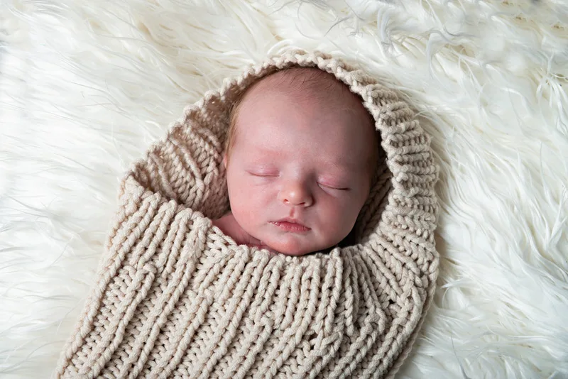Color photo of a newborn baby boy in a beige knit sack on a white rug. The photo is by Laura Pea Photography, a Long Island newborn and pet photographer. 