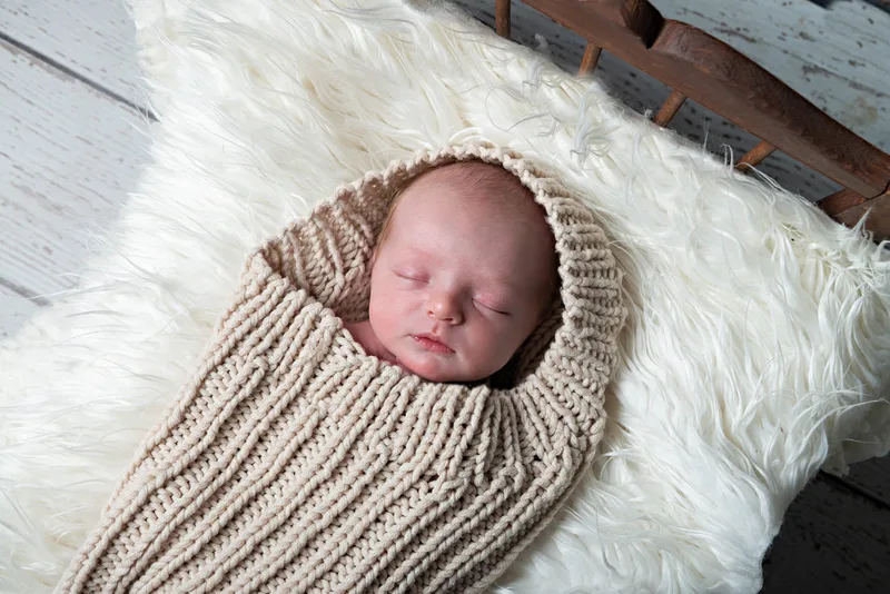 Color photo of a newborn baby boy in a beige knit sack on a white rug. The photo is by Laura Pea Photography, a Long Island newborn and pet photographer. 